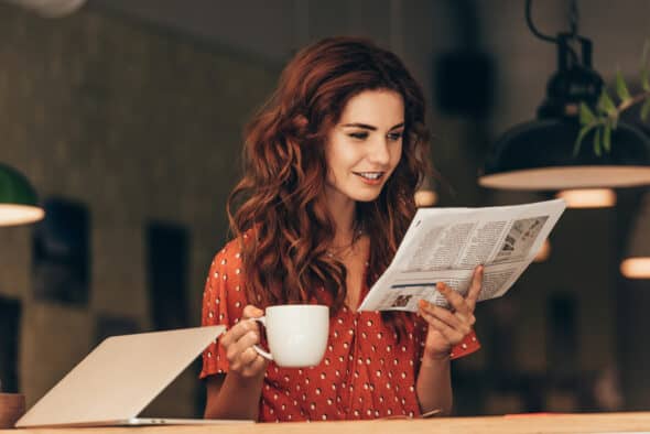 Woman with cup of coffee reading newspaper at table with laptop in cafe